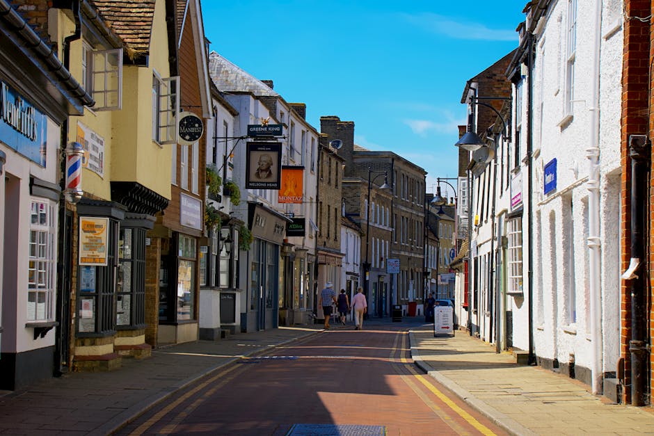 A narrow street on a bright, sunny day with clear blue skies, featuring a variety of small brick and plaster-fronted shops and cafés with signs hanging above the entrances. The pavement is lined with