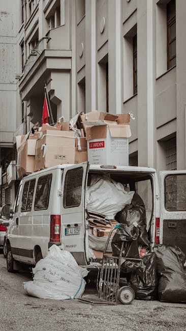 A white commercial van parked adjacent to a multi-storey building on a city street, with its rear doors open revealing an interior filled with stacked cardboard boxes, black and white plastic bags, an